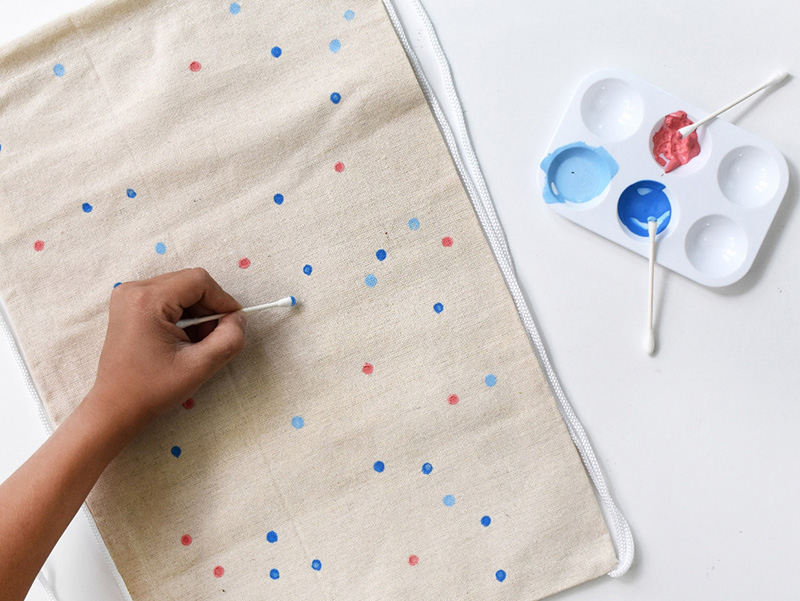 Using a qtip to paint blue and pink polka dots on a canvas tote